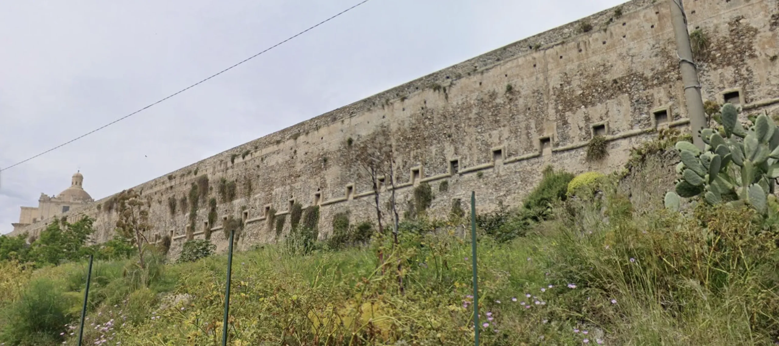Parete fortificata del castello vista da sotto, con vegetazione e feritoie spagnole